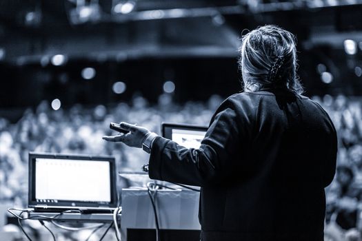 Female speaker giving a talk on corporate business conference. Unrecognizable people in audience at conference hall. Business and Entrepreneurship event. Black and white, blue toned image.