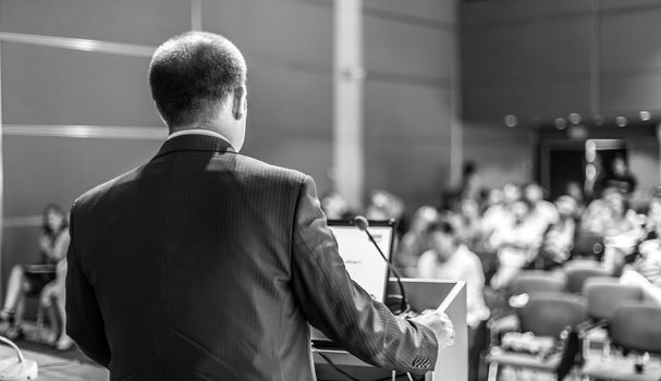 Speaker giving a talk on corporate business conference. Unrecognizable people in audience at conference hall. Business and Entrepreneurship event. Black and white image.