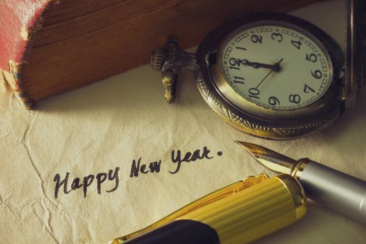 Vintage brass pen writing " Happy new year " on old paper with pocket watch and old book stacked on wooden table.