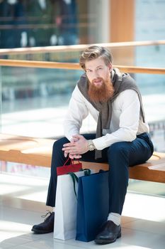 Sale and people - young bearded man with shopping bags sitting in shopping mall