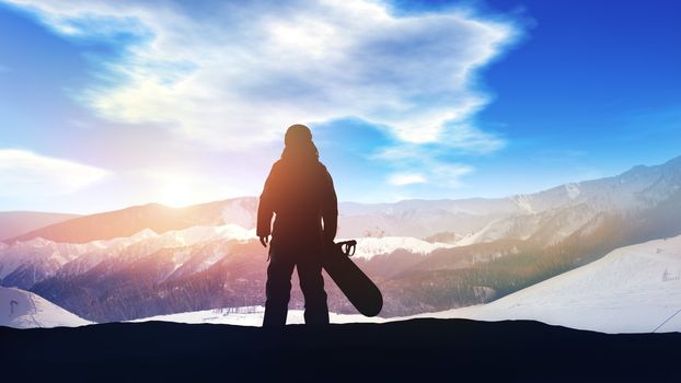 Against the snowy peaks of the mountains stands a dark silhouette of a snowboarder.