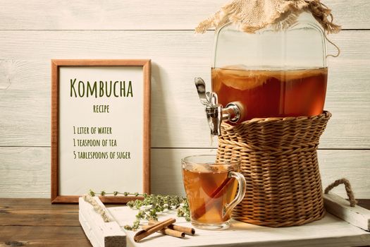 Fresh homemade kombucha fermented tea drink in a jar with faucet and in a cup on a white tray, the recipe for cooking in the frame, wooden background.