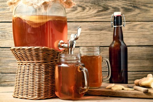 Fresh homemade Kombucha fermented tea drink in jar with faucet and in cans-mugs on wooden background.