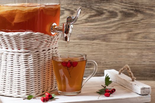 Fresh homemade kombucha fermented tea drink in a jar with faucet and in a cup on a white tray with hawthorn berrieson a wooden background, copyspace.