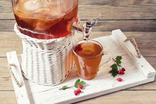 Fresh homemade kombucha fermented tea drink in a jar with faucet and in a cup on a white tray with hawthorn berrieson a wooden background.