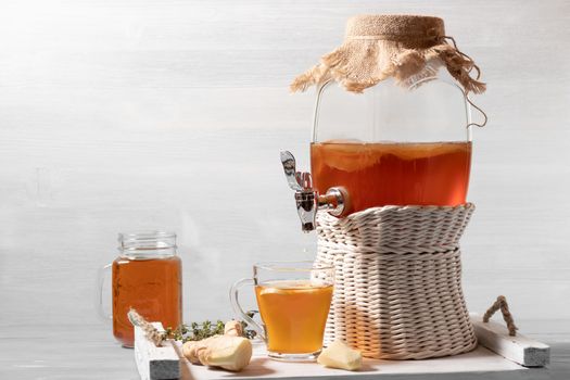 Fresh homemade kombucha fermented tea drink in a jar with faucet and in a cup and in mug on a white tray on a wooden background, copyspace.