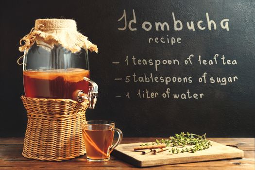 Fresh homemade kombucha fermented tea drink in a jar with faucet and in a cup and recipe on a dark wall.