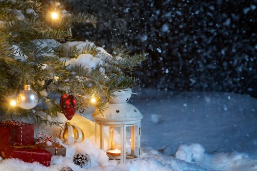 Christmas composition - gifts and a lantern in the snow under a Christmas tree decorated with lights and Christmas-tree decorations, copy space, place for text.