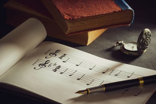 Music notes and old book with pocket watch on wooden table background in morning light. Writing chords by vintage pen. Closeup and copy space for text. Concept of Music lovers.