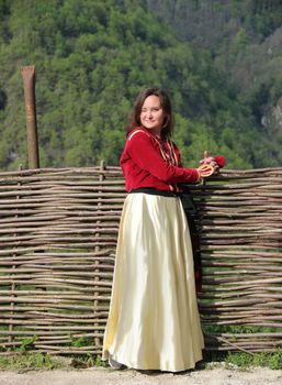 Photo of young brunette woman in a traditional georgian costume stands near a wicker fence on a background of mounains covered in a freen forest