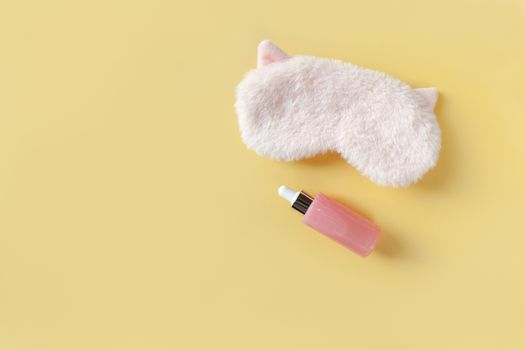 Pink fluffy fur sleep mask with small ears and bottle with serum for face on pastel yellow paper background. Top view. Concept of vivid dreams and skin care. Minimal style, copy space. Horizontal.