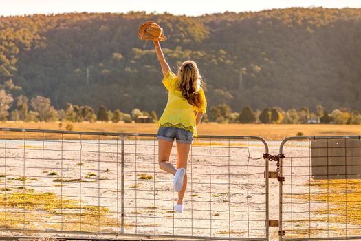 Aussie woman standing on a rural farm gate with one arm outstretched high above her head holding her hat