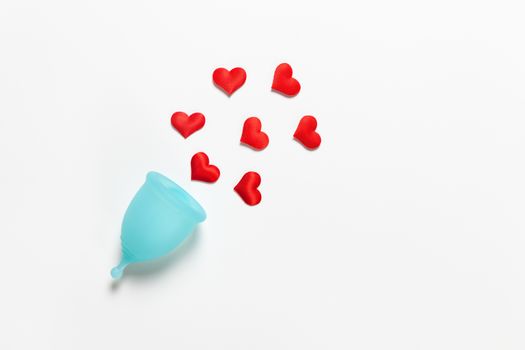 Turquoise menstrual cup on white background with red hearts. Concept zero waste, savings, minimalism, these days. Feminine hygiene product, flat lay, copy space. Horizontal.