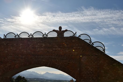 A silhouette of a person relaxing on a little bridge, with in the background the sun and mountains below the arc.