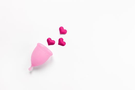 Pink menstrual cup on white background with crimson hearts. Concept zero waste, savings, minimalism, these days. Feminine hygiene product, flat lay, copy space. Horizontal.