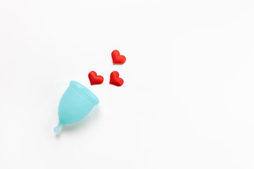 Turquoise menstrual cup on white background with red hearts. Concept zero waste, savings, minimalism, these days. Feminine hygiene product, flat lay, copy space. Horizontal.