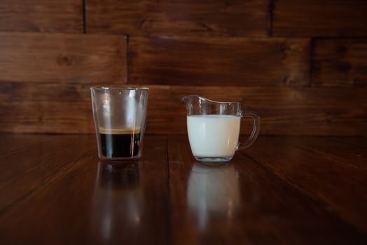 milk in glass milk jug with a cup of coffee in a transparent glass on wooden background