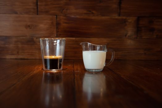 milk in glass milk jug with a cup of coffee in a transparent glass on wooden background