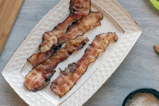 Fried bacon on white plate with cup of coffee, milk jug and toast. Breakfast on gray table