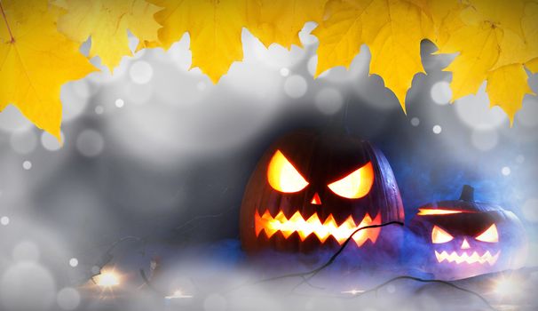 Halloween pumpkins head jack o lantern, candles and dry maple leaves in mist , bokeh lights and autumn yellow maple leaves frame