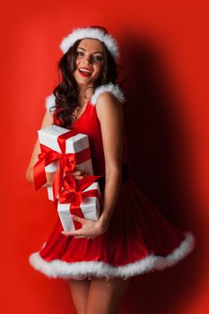 Pretty Pin-up style Santa girl in red hat and dress holding stack of gifts on red background