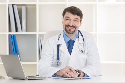 Male doctor working at office desk and smiling at camera , office interior on background