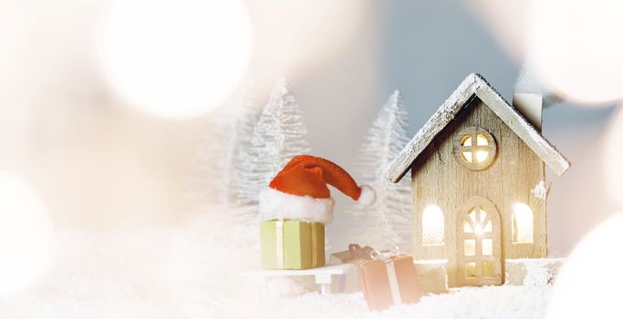 Christmas card with cozy small decorative house , gifts and Santa Claus cap on sleigh on snow over fairy tale winter fir forest background and falling snow . New Year design postcard . Copy space for text