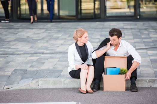 Fired business man sitting frustrated and upset on the street near office building with box of his belongings. He lost work. Business woman comforts and encourages him