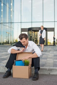 Fired business man sitting frustrated and upset on the street near office building with box of his belongings. He lost work