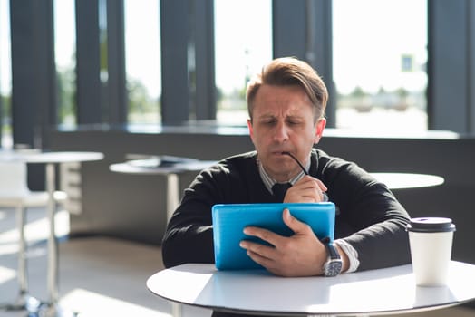 Portrait of thoughtful worried man sitting in cafe with digital tablet and biting glasses