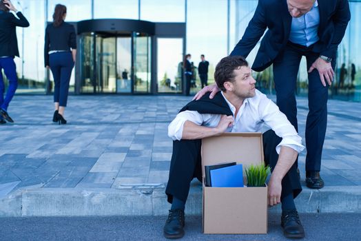 Fired business man sitting frustrated and upset on the street near office building with box of his belongings. He lost work. Business man comforts and encourages him