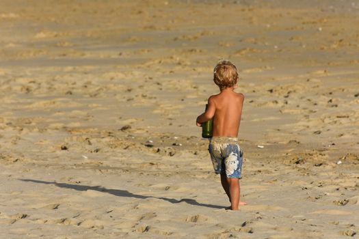 A curious blonde boy marching on sandy beach with an empty beer bottle as a trophy and pockets full of beach treasure, Mossel bay, South Africa