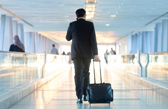 Rear view of unrecognizable formaly dressed businessman walking and wheeling a trolley suitcase at the lobby, talking on a mobile phone. Business travel concept.