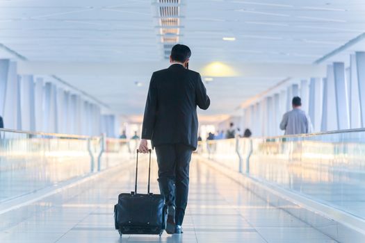 Rear view of unrecognizable formaly dressed businessman walking and wheeling a trolley suitcase at the lobby, talking on a mobile phone. Business travel concept.