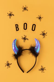 Halloween holiday flat lay. Party accessory, rim with devil horns, spiders and inscription Boo on orange background. Minimal style. Vertical. Trick-or-treat concept.