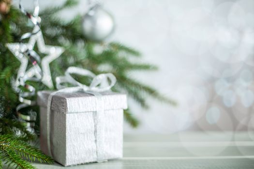 Silver Christmas new year gift with curly streamer ribbon bow over glittering bokeh lights background under fir tree with baubles