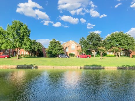 Riverside residential neighborhood in suburbs Dallas, Texas, USA. Row of single family detached homes with parked car on street, mature oak trees, high stone retaining wall, green grass lawn, blue sky