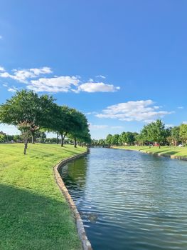 Waterfront subdivision in suburbs Dallas, Texas, USA. Clean river with high stone retaining wall and row of mature oak, bold cypress tree, green grass lawn along the banks. Single family detached home