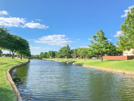 Waterfront subdivision in suburbs Dallas, Texas, USA. Clean river with high stone retaining wall and row of mature oak, bold cypress tree, green grass lawn along the banks. Single family detached home