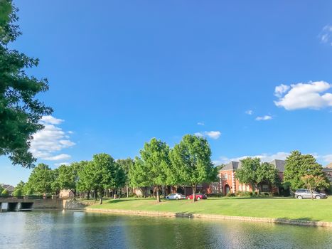 Riverside residential neighborhood in suburbs Dallas, Texas, USA. Row of single family detached homes with parked car on street, mature oak trees, high stone retaining wall, green grass lawn, blue sky