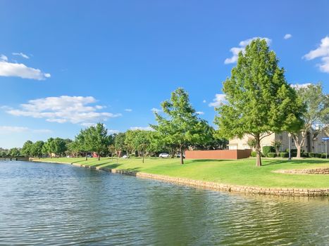 Beautiful waterfront house with row of mature bold cypress trees in suburbs Dallas, Texas, USA. Suburban single family detached home along river with high stone retaining wall, green grass lawn