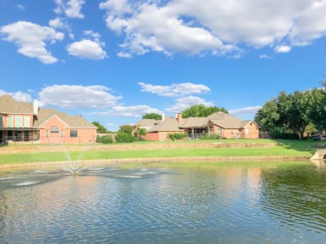 Lakefront houses with water fountain and green grass lawn bank in suburbs Dallas, Texas, USA. Suburban single family detached home along river with high stone retaining wall, sewage, mature tree