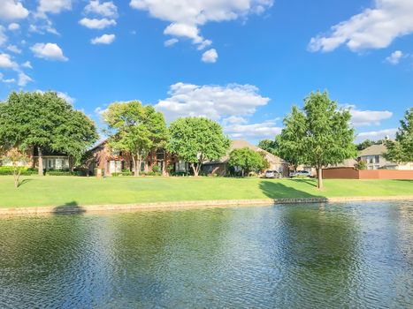 Beautiful waterfront house with row of mature bold cypress trees in suburbs Dallas, Texas, USA. Suburban single family detached home along river with high stone retaining wall, green grass lawn