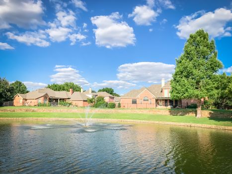 Lakefront houses with water fountain and green grass lawn bank in suburbs Dallas, Texas, USA. Suburban single family detached home along river with high stone retaining wall, sewage, mature tree