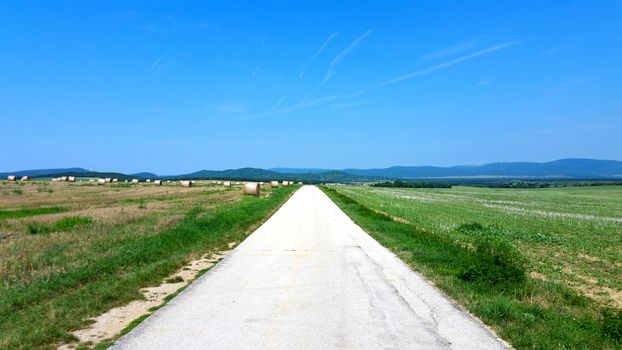 Light village road between hay rolls on wheat field in late summer on the blue sky background.