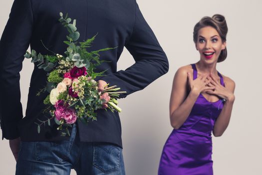 Man brings flowers to woman and hiding them behind back, womanin evening dress is surprised and happy