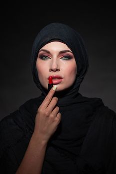 Portrait of an arab saudi emirates woman painting her lips with a lipstick on black background