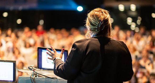 Female speaker giving a talk on corporate business conference. Unrecognizable people in audience at conference hall. Business and Entrepreneurship event.