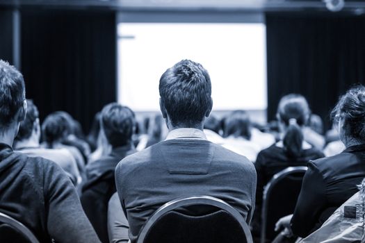 Business and entrepreneurship symposium. Speaker giving a talk at business meeting. Audience in conference hall. Rear view of unrecognized participant in audience.