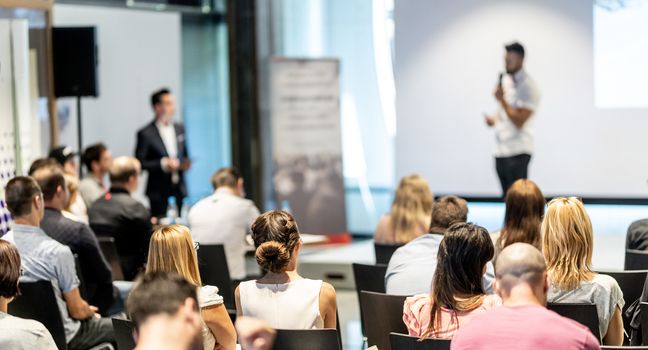 Business and entrepreneurship symposium. Speaker giving a talk at business meeting. Audience in conference hall. Rear view of unrecognized participant in audience.